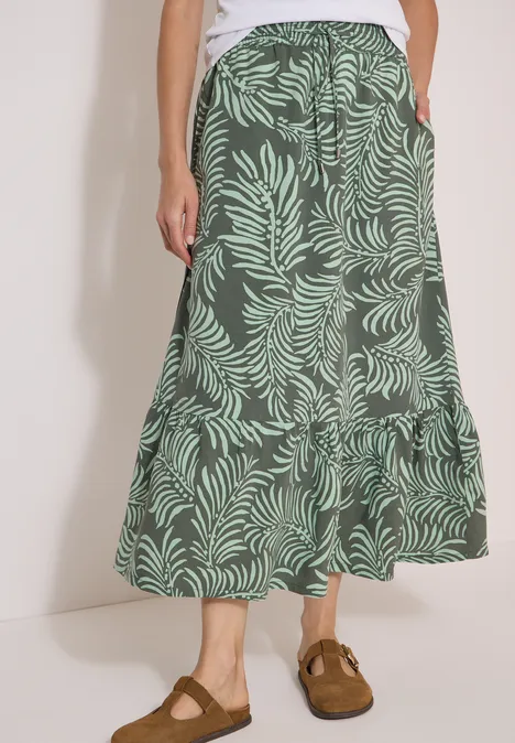 Maxirok met bladmotief khaki Persoon draagt een wit topje en een lange rok met groen bladerenpatroon, bruine klompen, handen in de zakken.