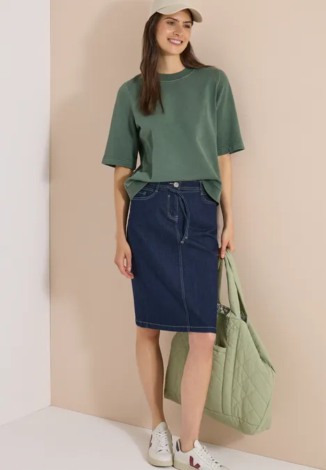 Jupe en jean arrivant aux genoux avec une ceinture rinsed wash Une femme porte un t-shirt vert, une jupe en jean bleu foncé, des baskets blanches et un sac matelassé vert.