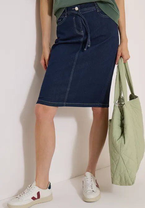 Jupe en jean arrivant aux genoux avec une ceinture rinsed wash Femme portant une jupe en jean, baskets blanches et tenant un sac cabas vert pâle.