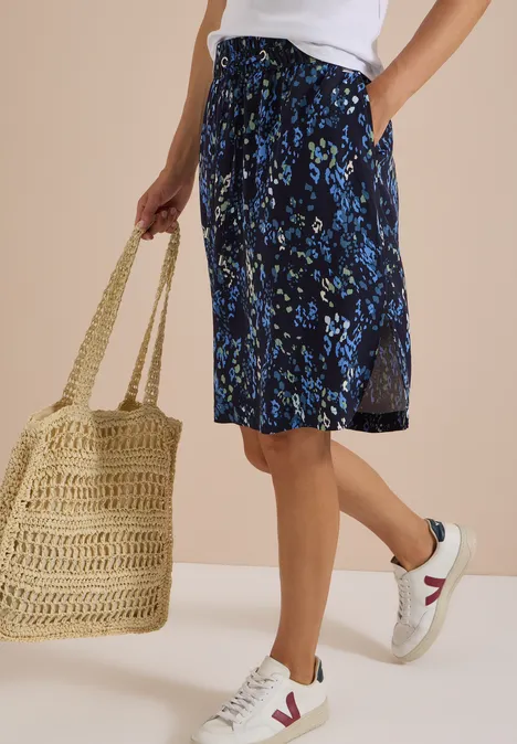 Dame in weißem Top, geblümtem blauem Midirock, weißen Sneakern und Korbtasche vor beigem Hintergrund.