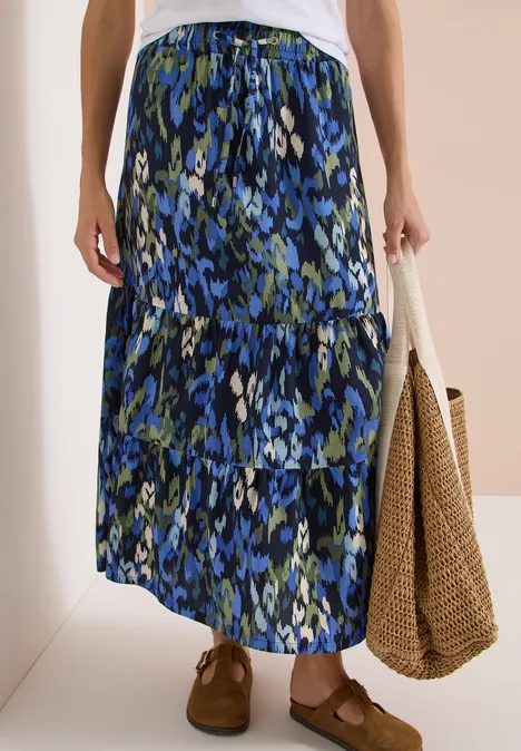 Maxirock mit Volants und Print urban dark blue Langer Damenrock in Stufenoptik mit blau-grünem Blumenmuster, kombiniert mit geflochtener Tasche und braunen Schuhen.
