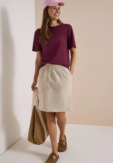 Vrouw met paars T-shirt, beige rok, bruine mocassins en strooien tas voor neutrale achtergrond.
