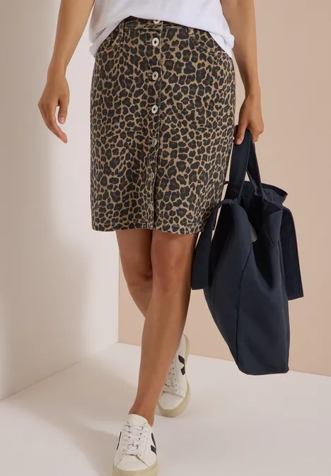Une personne porte une jupe au motif léopard jusqu'aux genoux, des baskets blanches et un sac bleu marine.