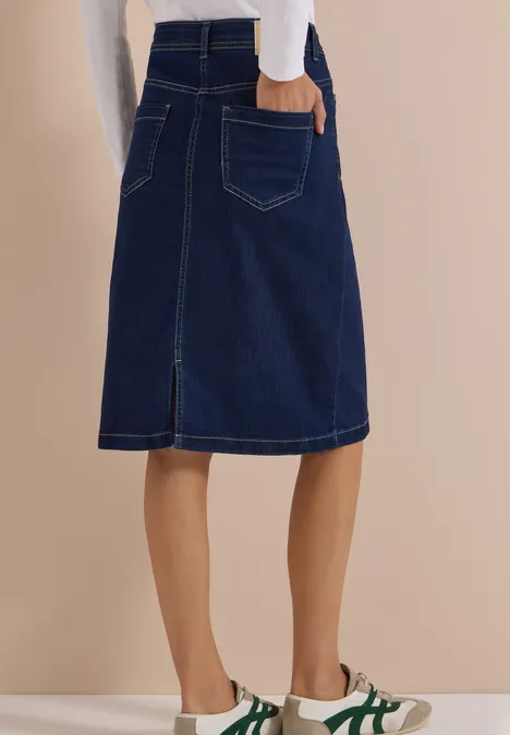 Donkerblauwe knielange denimrok met split achter, gecombineerd met witte sneakers met groene strepen.