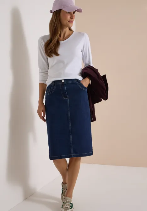 Vrouw in wit langarmshirt, knielange spijkerrok, beige sneakers en roze pet, met een bordeaux jasje in de hand.