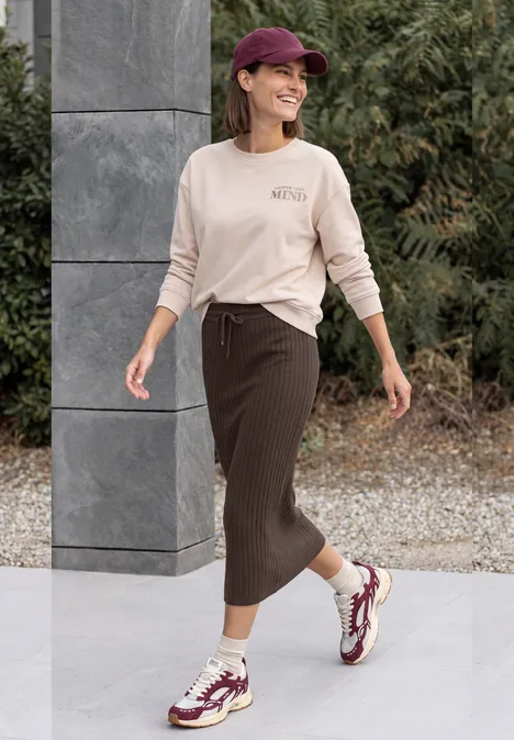 Eine lächelnde Frau spaziert draußen in einem lässigen Outfit mit Pullover, langer Rock, Cap und Sneakern.