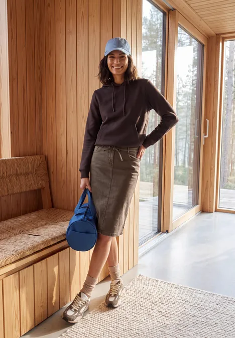 Vrouw in hoodie, rok en sandalen met blauwe tas poseert in lichte houten kamer bij een raam.