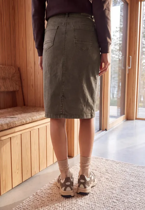 Een persoon staat in een lichte houten kamer, draagt een groene knielange rok en sneakers op een vloerkleed.