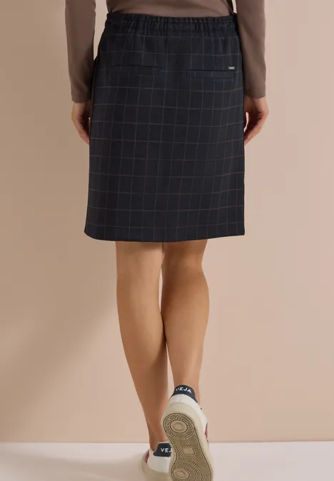 Achteraanzicht van een persoon in een geruite knie-lange rok, lang shirt en sneakers.