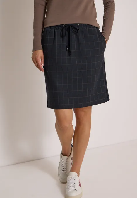 Vrouw in een knielange, zwart geruite rok met koord, beige longsleeve en witte sneakers.