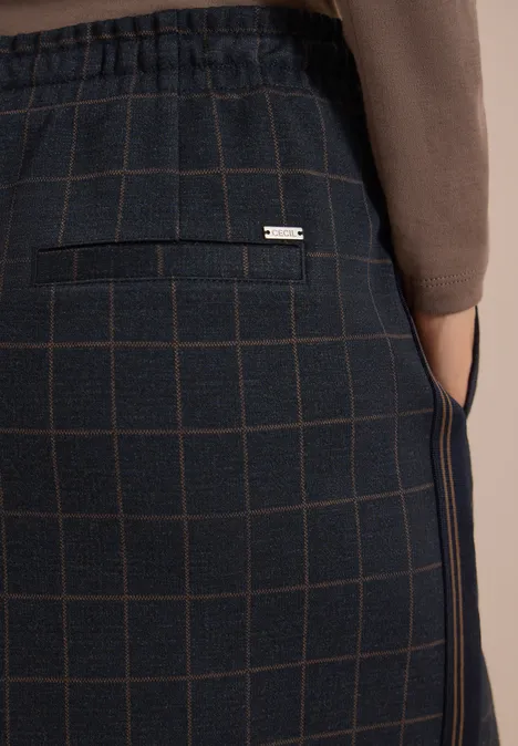 Close-up van donker geruit broek met achterzak en zilverkleurig label, gedragen met een bruin vest.