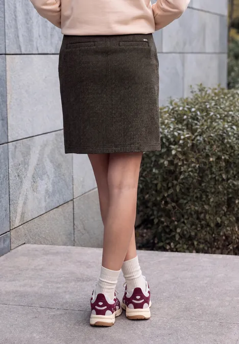 Van heup tot voeten: persoon in een bruine rok, witte sokken en witte sneakers met bordeaux details, voor een muur en struik.