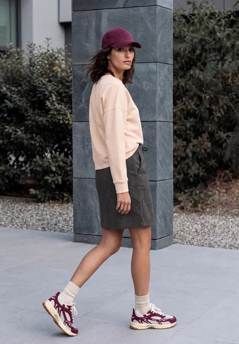 Vrouw in beige sweater, grijze rok, paarse sneakers en pet loopt buiten langs een zuil.