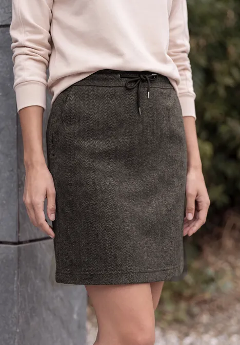 Iemand draagt een beige trui en een donkergroene wollen rok, staand naast een betonnen muur.