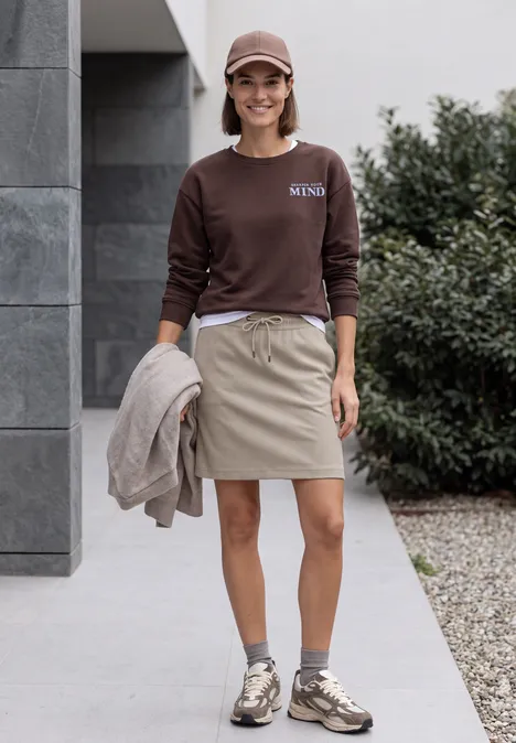 Vrouw in bruine trui en beige rokje met sneakers, jas in de hand, glimlachend voor een modern huis.
