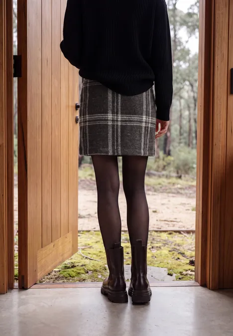 Vrouw staat in een houten deurkozijn, draagt een geruite rok, zwarte panty en enkellaarzen, uitzicht op bos.