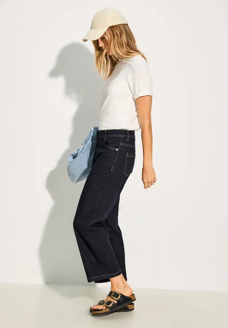 Frau mit weißem Shirt, dunkler Jeans, Kappe und Sandalen, trägt eine blaue Jacke.