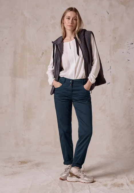 Femme en blouse blanche, gilet et pantalon bleu, debout devant un mur clair.