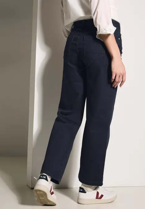 Vrouw in donkerblauwe jeans, wit shirt en witte sneakers staat bij een muur.