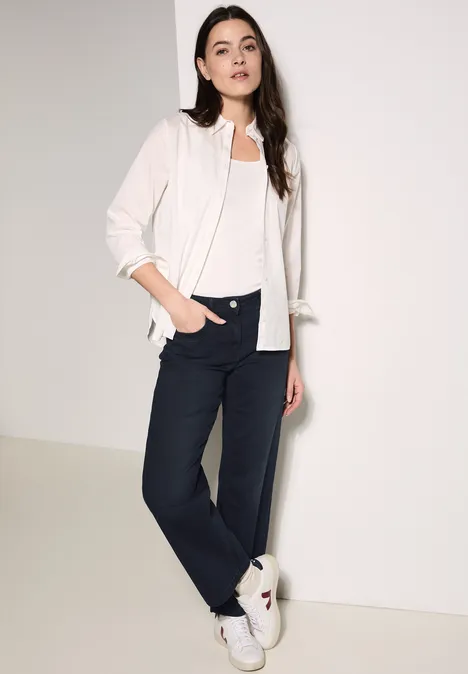 Vrouw met wit overhemd, wit shirt en donkere broek leunt relaxed tegen de muur.