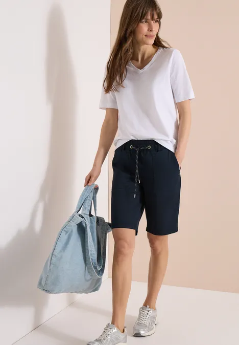 Femme en t-shirt blanc, bermuda bleu marine, baskets argentées et sac en jean, posée devant un mur beige et blanc.