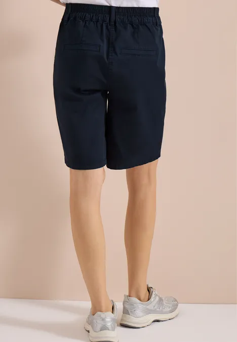 Vue de dos d'une personne portant un short bleu marine au genou et des baskets claires sur fond beige.