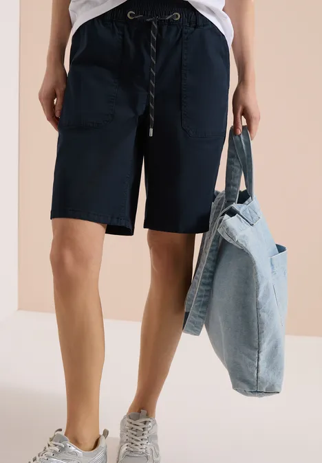 Person en short bleu marine, t-shirt blanc et baskets, tenant un sac en jean clair.