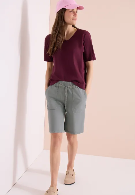 Persoon in bordeaux T-shirt, grijze bermuda en beige sneakers, leunend tegen een witte muur.