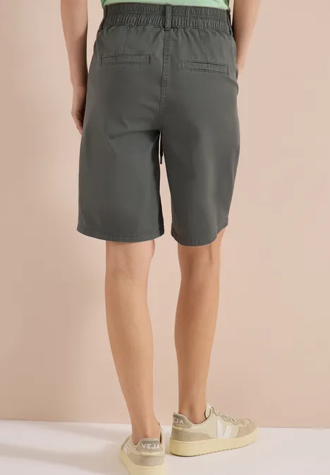 Person steht in grauen Shorts und hellen Sneakern, Rückenansicht vor beigem Hintergrund.