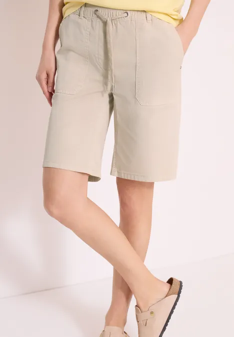 Personne porte un short beige jusqu'aux genoux avec poches et ceinture à nouer, associé à des mules claires.