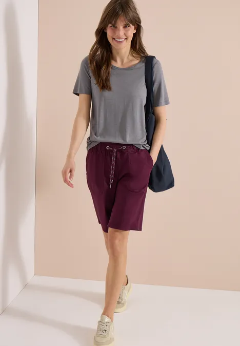 Une femme en t-shirt gris, bermuda bordeaux et baskets, portant un sac bandoulière bleu.