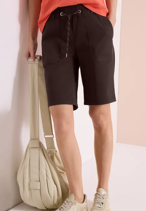 Une personne porte un short noir, des baskets claires et tient un sac beige.