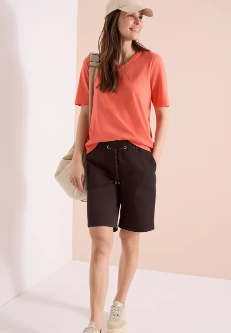 Femme en t-shirt corail, bermudas noirs, casquette crème et baskets, sac épaule, sur fond clair.