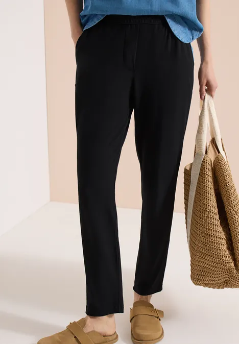 Personne porte un pantalon noir décontracté et des chaussons, tenant un sac tressé.