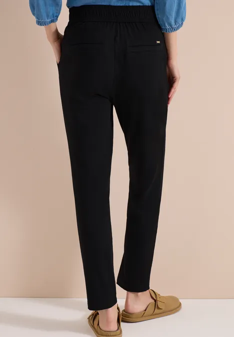 Personne vue de dos portant un pantalon noir, un haut bleu et des mocassins beiges sur fond beige.