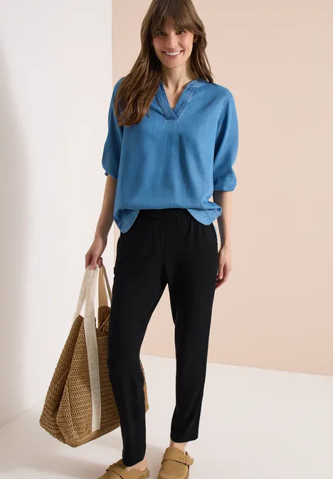 Femme en haut bleu ample à col V, pantalon noir et grand sac en paille tressée sur fond uni.