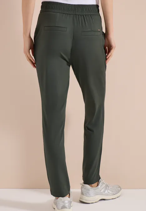 Vue arrière d'une personne portant un pantalon de jogging étroit foncé et des baskets claires sur fond beige.