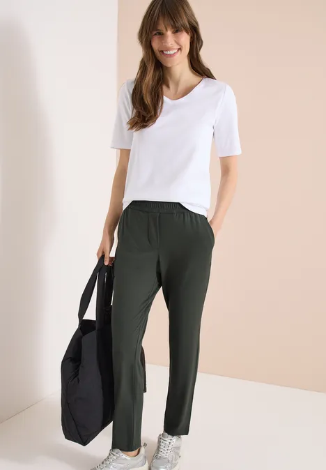 Femme portant un t-shirt blanc, un pantalon vert foncé et tenant un sac noir.