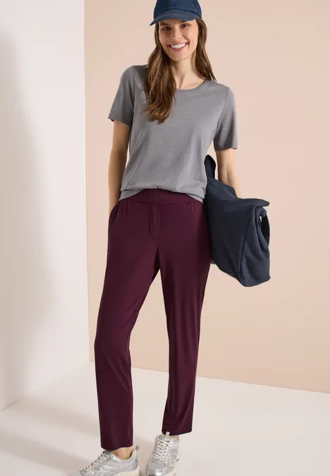 Femme en t-shirt gris, pantalon bordeaux et baskets, sac bleu à la main, style décontracté.