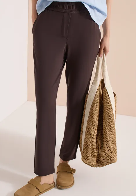 Personne en pantalon marron foncé décontracté à taille élastique, chaussons marron et sac tressé à la main.