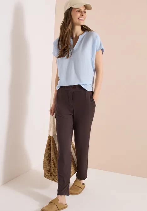 Femme en t-shirt bleu, pantalon brun, sandales plates et casquette, tenant un sac en paille devant fond beige.