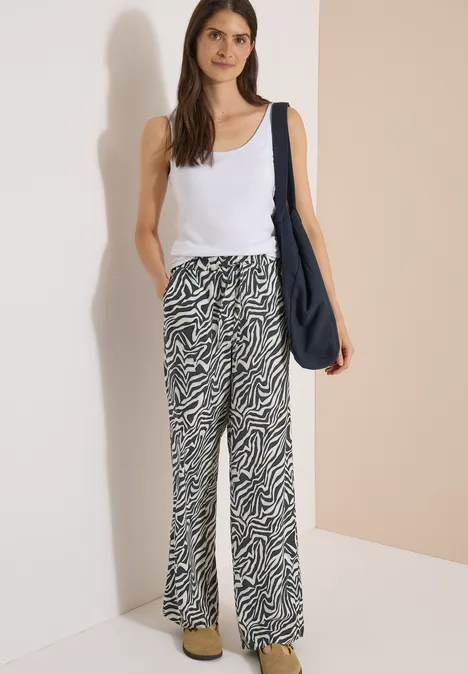 Femme en débardeur blanc, pantalon large à motif zébré et sac bandoulière bleu marine, debout devant un mur beige.