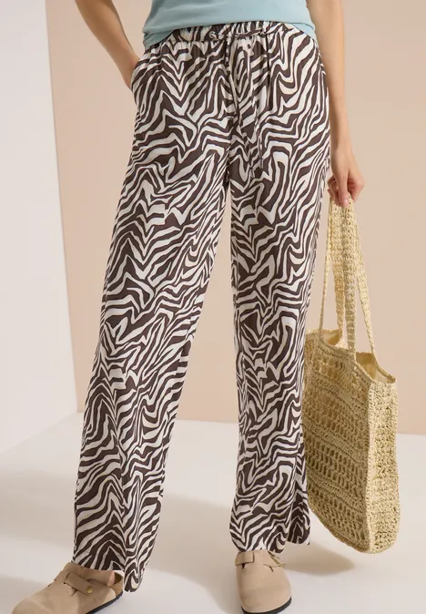 Femme porte un pantalon large imprimé zèbre noir et blanc, un top clair, un sac en osier et des chaussures plates.
