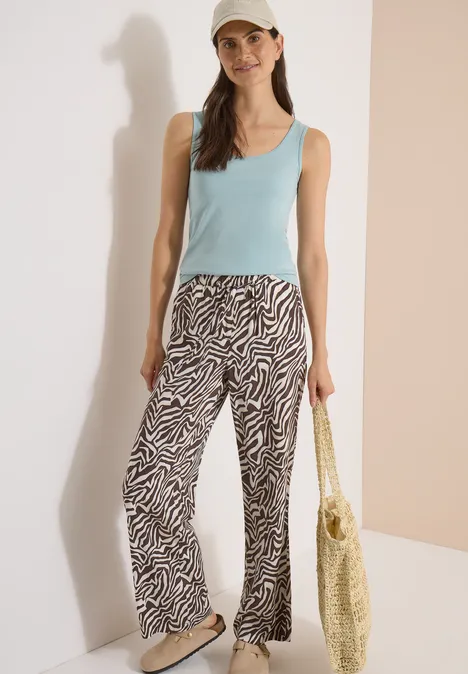 Femme en débardeur bleu clair, pantalon zèbre et casquette, portant un sac tressé devant un fond neutre.