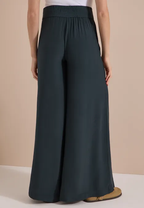 High Waist Wide Leg Hose im Loose Fit palm breeze green Person von hinten in weiten, dunklen, fließenden Hosen.