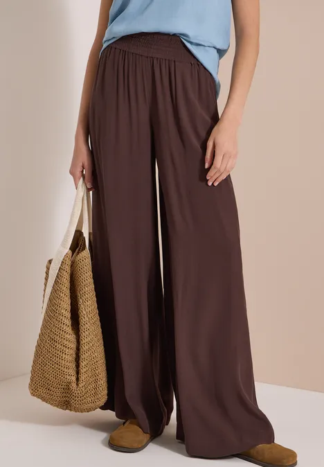 High Waist Wide Leg Hose im Loose Fit brown cacao Frau in weiter brauner Hose, hellblauem Top, Korbtasche und Mules.