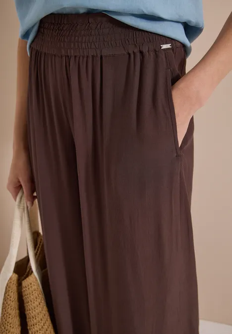 High Waist Wide Leg Hose im Loose Fit brown cacao Nahaufnahme einer braunen, locker sitzenden Hose mit Gummibund und Seitentaschen, Hand in Tasche, Strohtasche am Arm.
