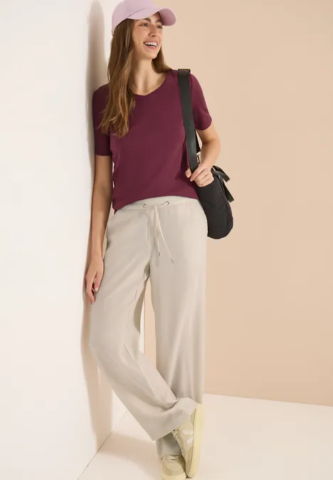 High Waist Straight Leg Hose im Loose Fit grain beige Frau im lässigen Outfit: bordeauxrotes T-Shirt, helle weite Hose, gelbe Sneaker, schwarze Tasche und rosa Cap.