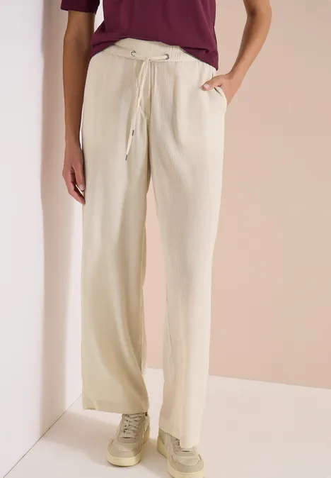 High Waist Straight Leg Hose im Loose Fit grain beige Person trägt beige, weite Hose mit Kordelzug und Taschen, kombiniert mit einem bordeauxroten Oberteil und Sneakern.