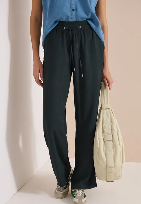 High Waist straight legs broek in Loose Fit palm breeze green Zwarte, wijde broek met trekkoord, blauw topje, houdt beige doorgestikte jas vast, draagt sneakers.